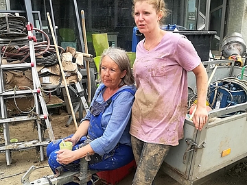 Zwei Frauen in verschlammten Arbeitssachen und Gummistiefeln sitzen bzw. stehen an einem Anhänger vor einem zerstörten Haus und machen eine kleine Pause
